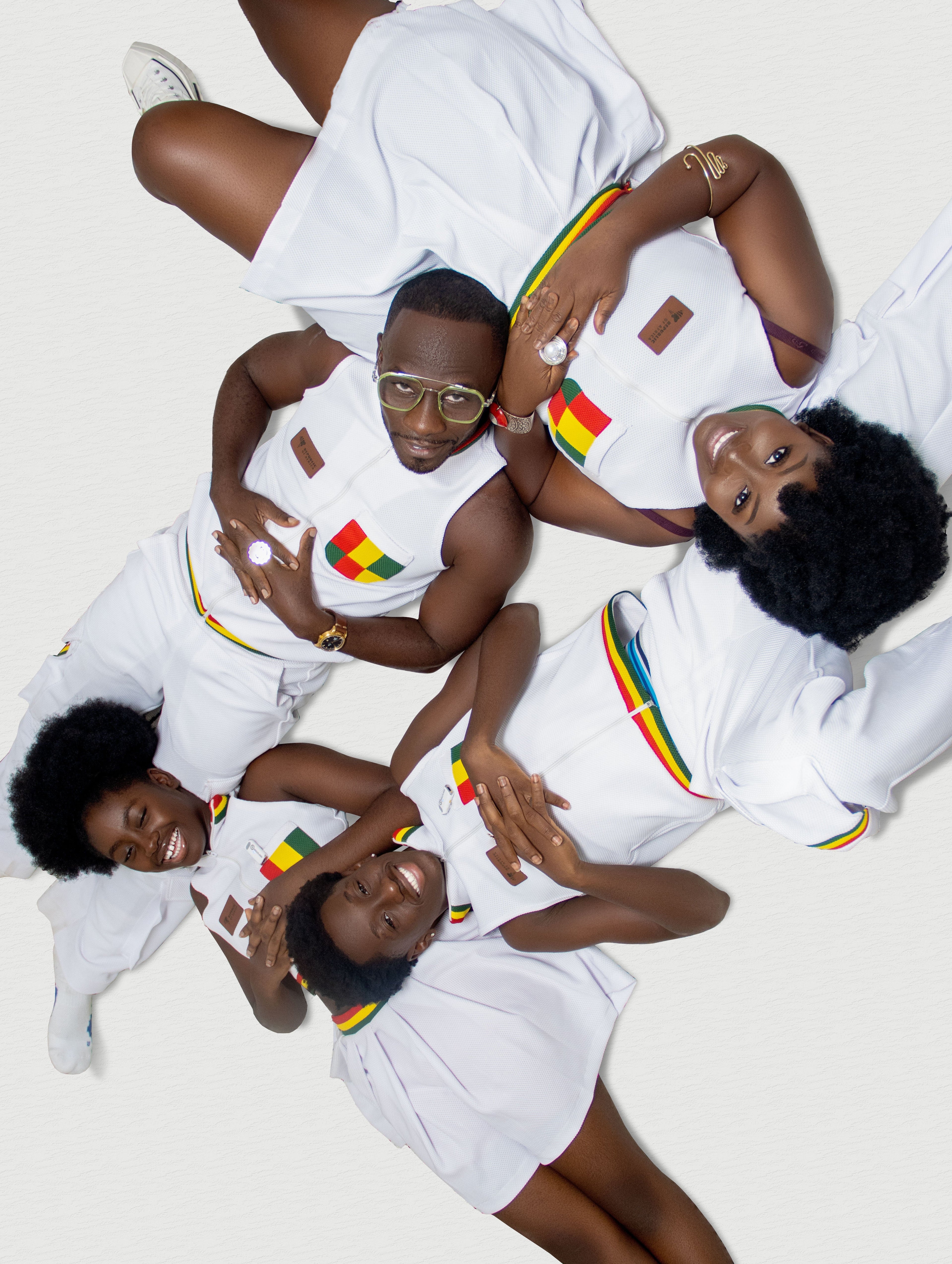 Okyeame Kwame and family lying on a white surface in matching white sleeveless jersey outifts, featuring red-gold-and-green pockets and trims.