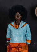 Woman with large afro wearing the blue Thabi crop top and orange pants, standing against a dark background.